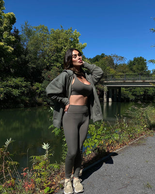 a woman standing on the side of a river wearing a green jacket and black leggings